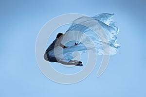 Portrait of flexible woman, graceful ballerina dancing with fabric, cloth isolated on blue studio background. Grace, art