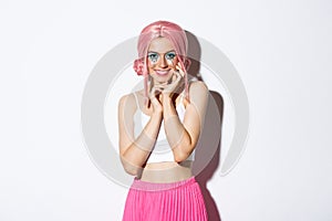 Portrait of flirty beautiful woman with pink wig, halloween makeup, smiling and looking coquettish, standing over white