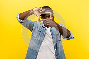 Portrait of focused curious man looking through photo frame made of hands. indoor studio shot isolated on yellow background