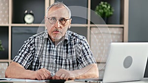 Portrait of focused elderly business male posing at modern workplace with laptop pc sitting at desk