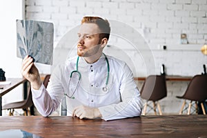 Portrait of focused male doctor in white uniform examining brain computerized tomography scan sitting at desk in