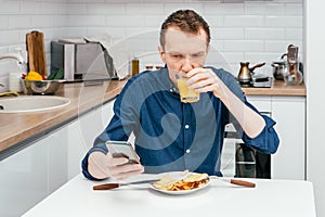 Portrait of focused middle-aged man in blue shirt looking at mobile phone drinking orange juice in white modern kitchen.