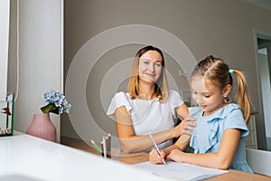 Portrait of focused primary daughter doing homework with young mother sitting at home table by window.