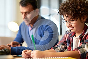 Portrait of focused teenaged latin boy in glasses sitting at the desk together with his father and doing homework at