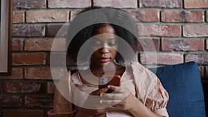 Portrait of focused young African-American woman using smartphone, typing message sitting by soft couch at home.