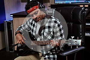 Portrait of focused young man, male artist in headphones playing guitar while sitting in recording studio