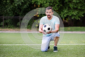 Portrait of football coach or gym teacher standing on one knee holding and giving soccer ball to camera and talking on green field