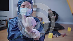 Portrait of forensic investigator in uniform putting on white gloves and looking at camera. Female professional working