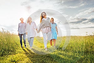 Portrait of four cheerful smiling women holding hand in hand walking by a high green grass meadow. They looking at the camera.