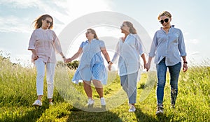 Portrait of four cheerful smiling women holding hand in hand walking by a high green grass meadow. They looking at the camera.