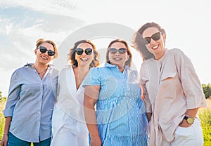 Portrait of four cheerful smiling women in sunglasses embracing and looking at the camera during outdoor walking. Woman\'s