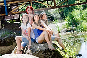 Portrait of four girls friends young women having fun sitting on stone posing & looking at camera on summer outdoors
