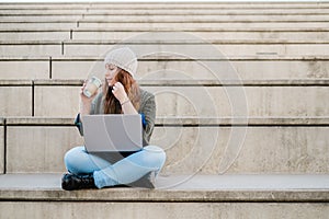 Portrait of freelancer woman using laptop and drinking coffee while sitting on stairs in city street. Blogger concept.