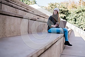 Portrait of freelancer woman using laptop and drinking coffee while sitting on stairs in city street. Blogger concept.