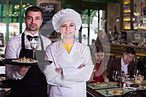 Portrait of frendly waiter and woman cook who are standing with tray