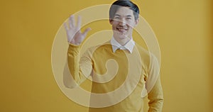 Portrait of friendly Asian man waving hands and smiling looking at camera on yellow background