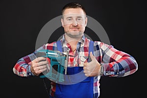 Portrait of a friendly carpenter with an electric jigsaw in his hand showing a thumb up. Studio shot on black background