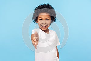 Portrait of friendly cheerful little boy with curly hair in white T-shirt giving hand to handshake and smiling at camera