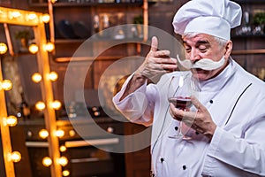 Portrait of friendly elderly male professional sommelier wearing apron and hat tasting red wine.