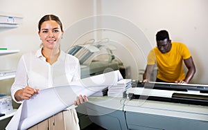 Portrait of friendly female employee of printing house with stack of notebooks in her hands