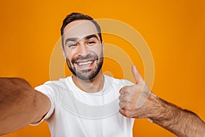 Portrait of friendly man 30s in t-shirt laughing while taking selfie photo,  over yellow background