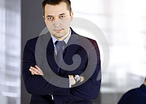 Portrait of a friendly middle aged businessman in a dark blue suit, with crossed arms, while standing in a sunny office