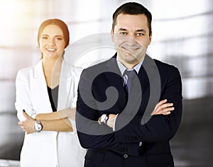 Portrait of a friendly middle aged businessman in a dark blue suit, standing with crossed arms together with a colleague