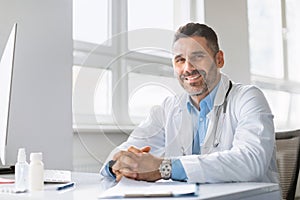 Portrait of friendly middle aged male doctor wearing medical workwear posing at clinic, sitting at workdesk