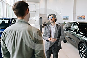 Portrait of friendly professional car dealer selling new automobile to unrecognizable male client explaining