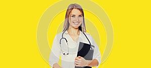 Portrait of friendly smiling young woman doctor with stethoscope and folder of documents on yellow background