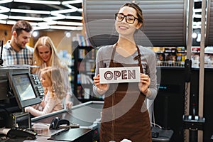 Portrait of friendly woman cashier holding open sign