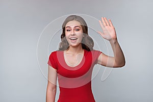 Portrait of friendly young brunette woman smiling and waving hand to say hello, say hi and welcome, goodbye gesture