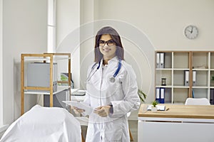 Portrait of friendly young female doctor, nurse or intern posing in white coat in medical office.