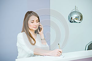 Portrait of friendly young woman talking on phone and making note behind the reception desk. Administrator in the office, clinic,
