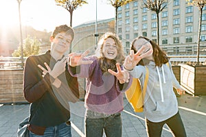 Portrait of friends teen boy and two girls smiling, making funny faces, showing victory sign in the street. City background,