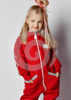 Portrait of frolic blonde kid girl in red overall jumpsuit and cool hairstyle standing with her hands in pockets