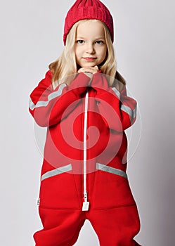 Portrait of frolic blonde kid girl in red overall jumpsuit and winter hat standing with her hands at her chin