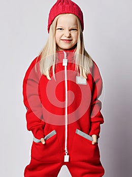 Portrait of frolic sly blonde kid girl in red overall jumpsuit and winter hat standing with her hands in pockets