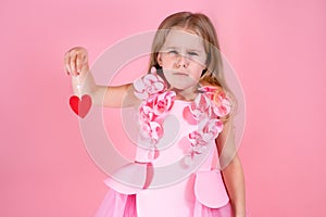 Portrait of frustrated cute little blue-eyed girl with long fair hair in pink dress holding red paper heart on string.
