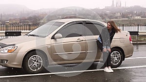 Portrait of a frustrated girl on an empty road in the rain near her broken car.