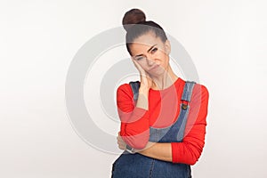 Portrait of frustrated indifferent girl with hair bun in denim overalls leaning on hand expressing disinterest