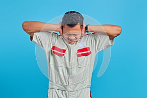 Portrait of frustrated young asian mechanic covering ears with palms over blue background