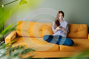 Portrait of frustrated young woman sitting on couch at home using smartphone looking away pondering of problem. Anxious