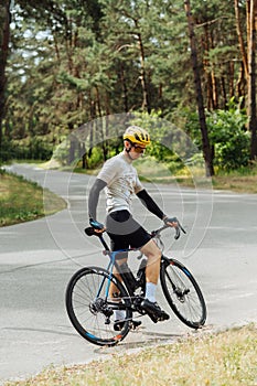 Portrait of a full-length man cycling in outfit standing with a bicycle outside the city in the woods on an asphalt road. Vertical