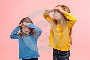 Portrait of funny amusing two little girls holding hands over eyes and looking far away with curious attentive expression