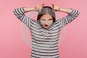 Portrait of funny angry woman in striped sweatshirt showing bull horn gesture and looking with hate rage