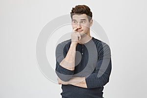 Portrait of funny athletic guy with stupid expression, sticking finger in nose and looking aside, standing against gray