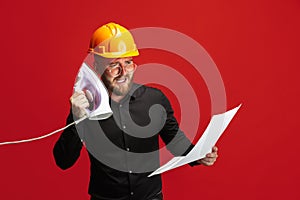 Portrait of funny bearded man, builder, designer or architect with professional equipment isolated on red studio