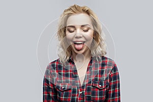 Portrait of funny beautiful blonde young woman in casual red checkered shirt standing with closed eyes and tongue out