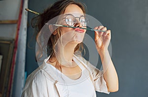 Portrait of a funny beautiful female artist in eyeglasses holding on her lips a painting brush in her workshop. Pretty young woman